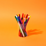 A clear plastic cup filled with assorted colorful pens, standing on a solid orange background.